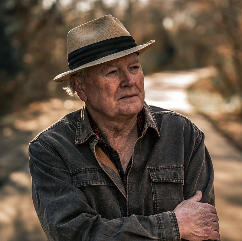 Alan Burton with a classy hat looking off to the distance while standing on a wooded path. About the author photo