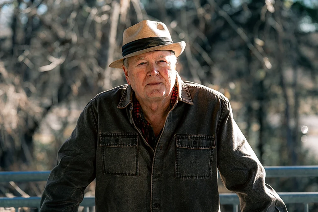 Alan Burton with a hat facing the camera in front of a wooded area. About the author photo